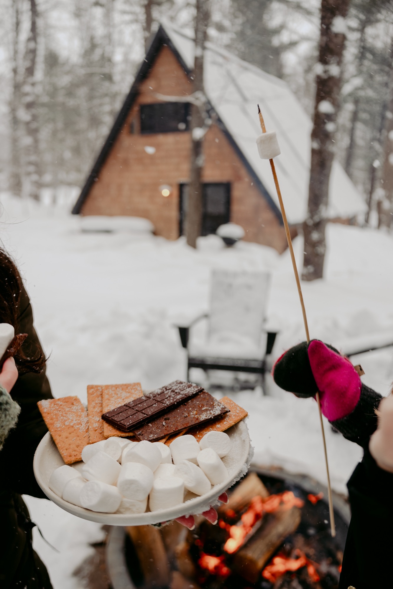 S'mores supplies and a roasting marshmallow with a snowy A-Frame cabin in the background