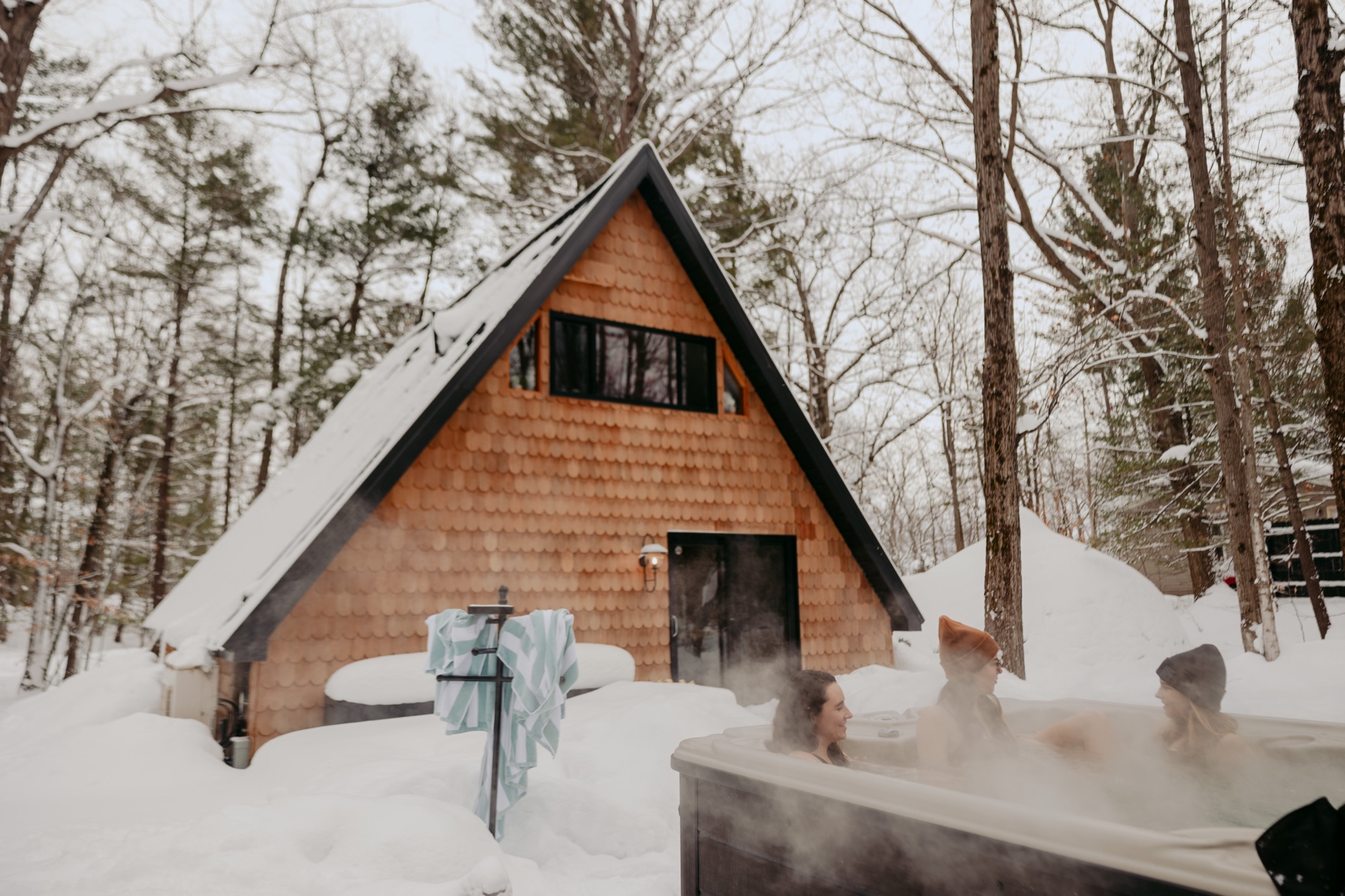Three friends relaxing in a steaming hot tub in front of the A-Frame cabin in winter