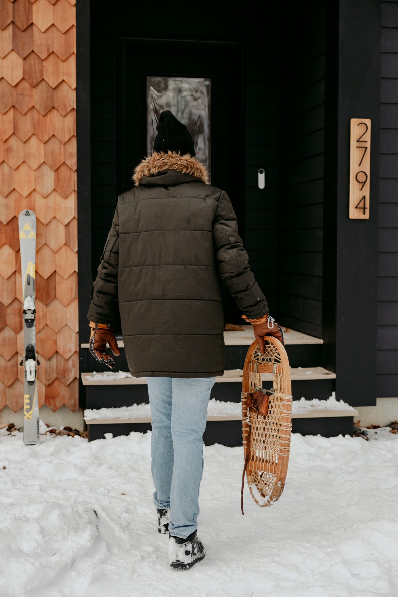 Person holding snowshoes in front of a modern cabin with skis propped against the wall